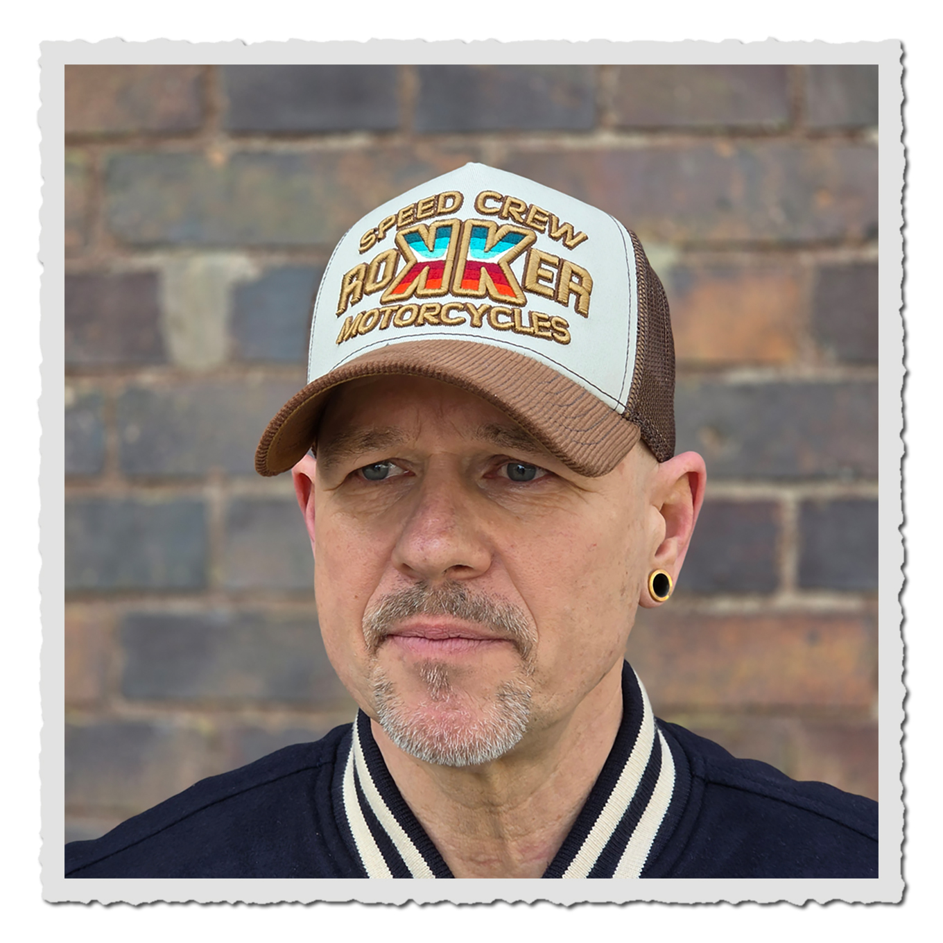 Nahaufnahme einer Person mit heller und brauner Cap vor Backsteinwand, Gesicht und Jackenkragen sichtbar.